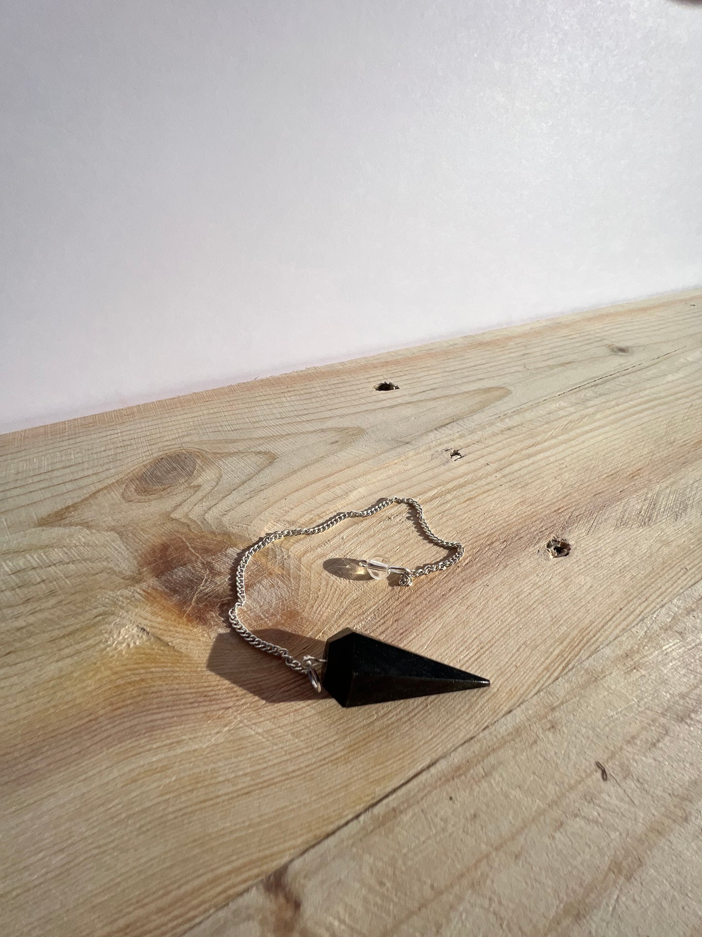 A zoomed out view of a black obsidian pendulum sitting on a pine board with a white backdrop.